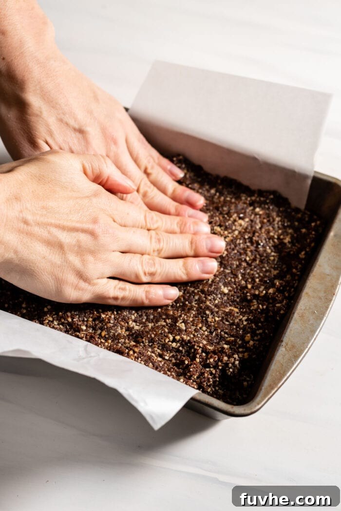 Hands pressing chocolate dough into a small square baking tray lined with parchment paper, creating an even layer.