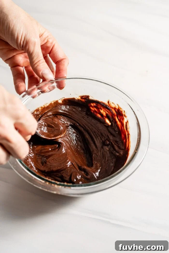 Hands using a spoon to mix a bowl of melted chocolate with coconut cream, making a smooth ganache.