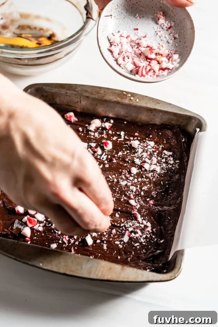 Hand sprinkling crushed candy canes over a layer of melted chocolate ganache on brownies in a pan.