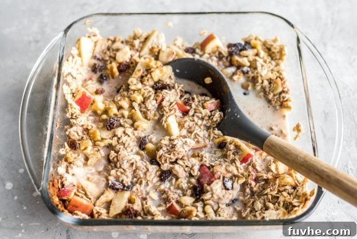 Mixed ingredients for apple cinnamon baked oatmeal in a glass baking dish with a mixing spoon resting in the dish, ready for baking.
