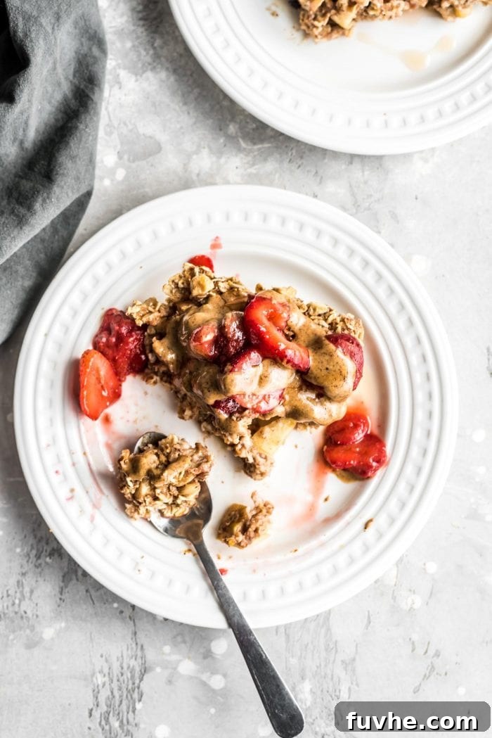 A square of baked apple oatmeal topped with almond butter and strawberries on a plate with a fork resting on it, inviting you to take a bite.