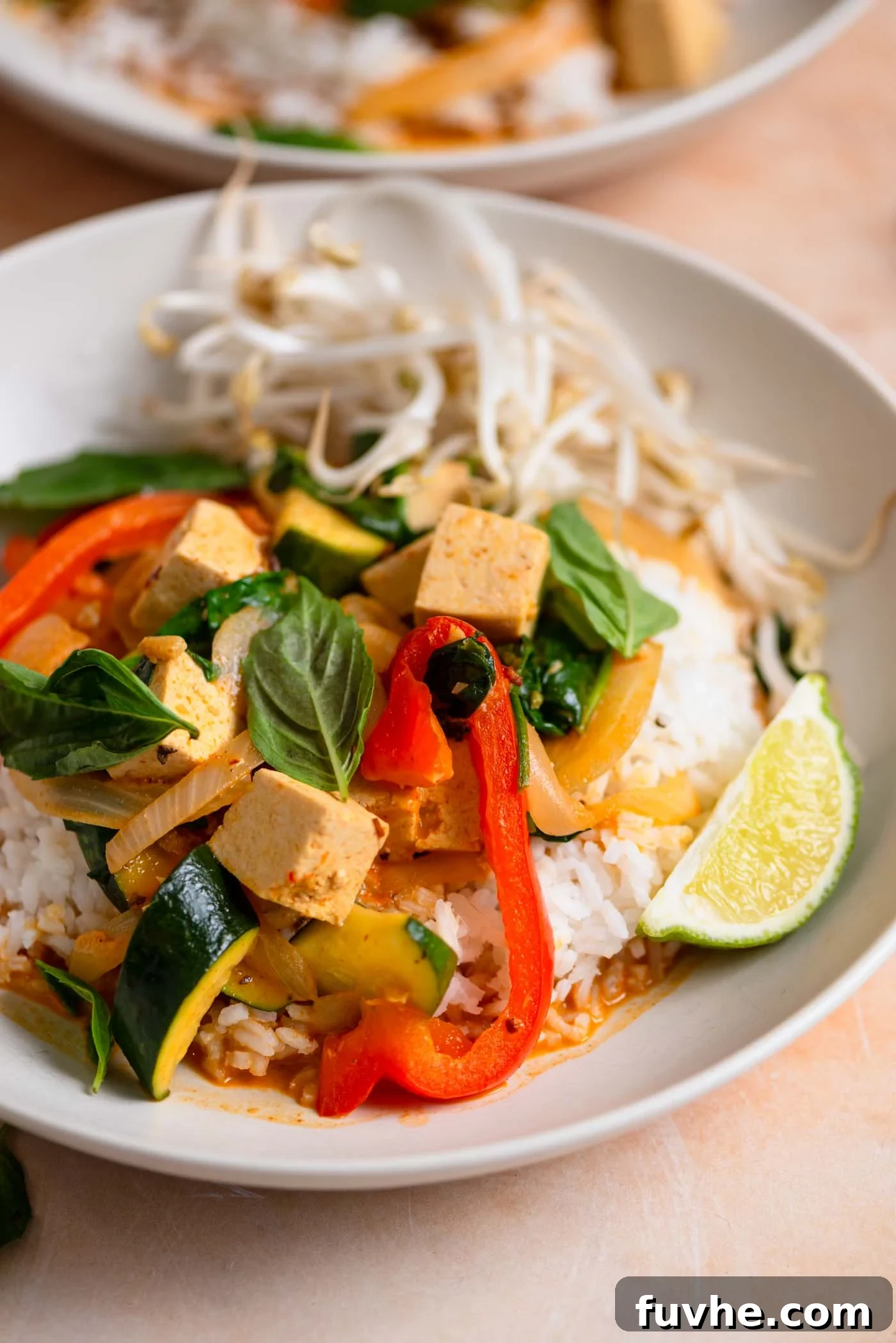 A vibrant bowl of vegan Thai red curry featuring perfectly cooked tofu cubes, tender bell peppers, and zucchini, garnished with fresh bean sprouts, aromatic basil leaves, and a bright lime wedge. The rich red curry sauce promises a burst of authentic Thai flavor.