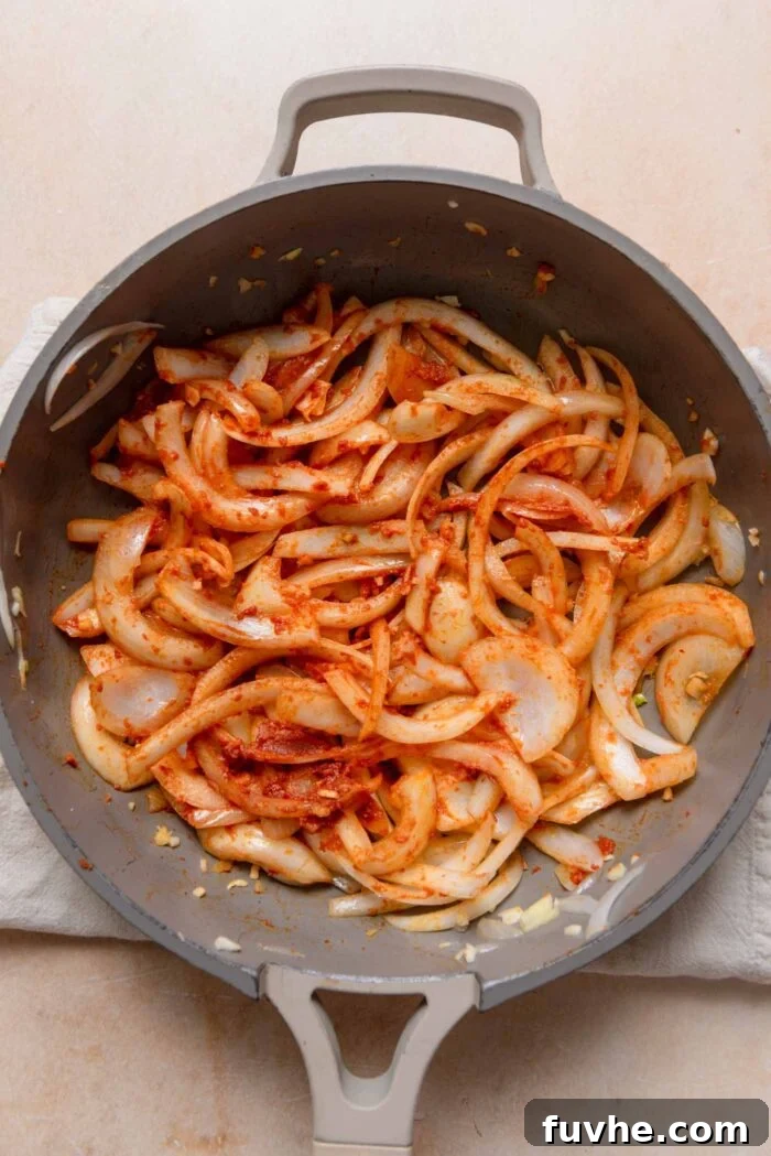 Sliced onions continuing to cook, now mixed with vibrant red curry paste, infusing the pan with rich Thai aromas.