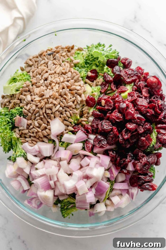 Diced red onion, dried cranberries, sunflower seeds and broccoli in a large glass mixing bowl.