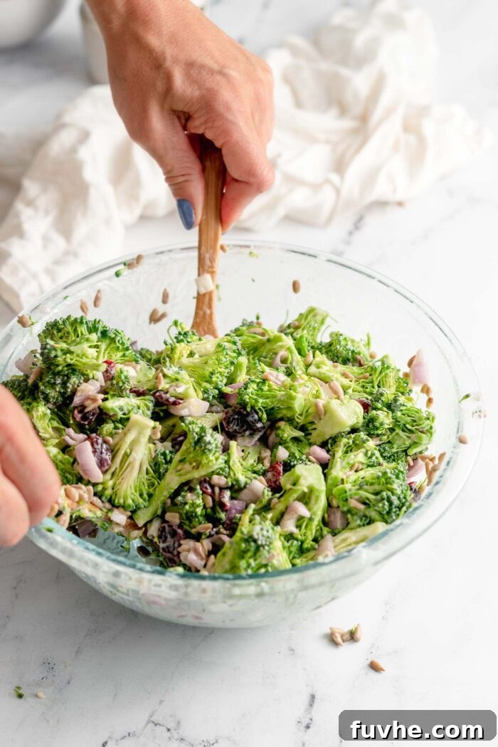 Hands using salad tongs to mix vegan broccoli salad in a large glass mixing bowl on a marble surface.
