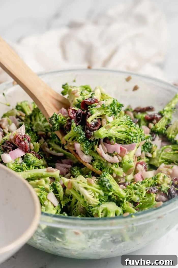 Wooden spoon scooping a serving of vegan broccoli salad with cranberries, red onion and sunflower seeds from a large glass mixing bowl into a small bowl.