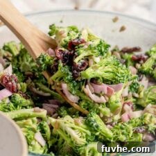 Wooden spoon scooping a serving of vegan broccoli salad with cranberries, red onion and sunflower seeds from a large glass mixing bowl into a small bowl.
