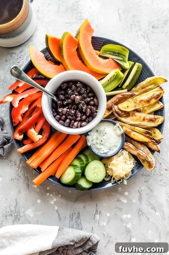 A plate with raw sliced veggies, roasted potato, black beans, papaya and a dip.
