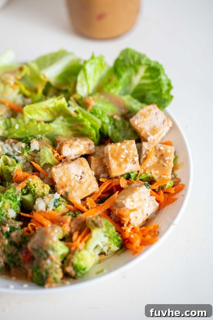 A close up of tofu in a bowl with lettuce, carrot, broccoli and peanut sauce.