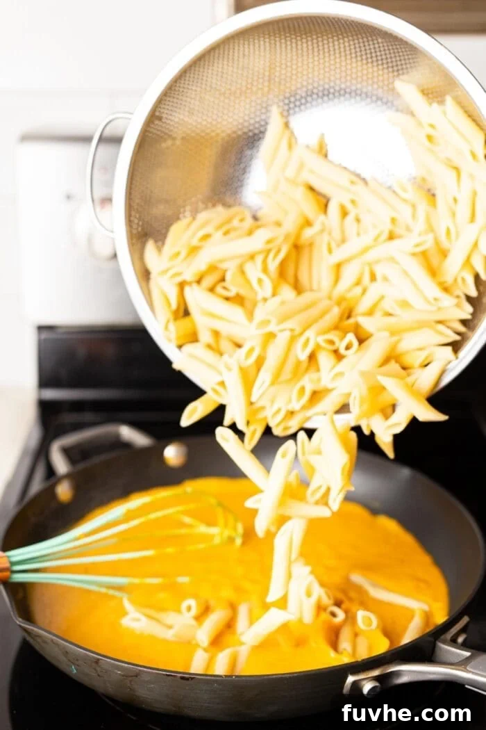 Silky Vegan Pumpkin Pasta 6 Dumping cooked penne pasta into a skillet of pumpkin cream sauce on a stovetop.