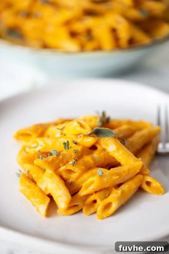 Silky Vegan Pumpkin Pasta 7 A plate of pumpkin penne pasta topped with bits of fresh sage on a small plate with a fork resting on it. A bowl of pumpkin pasta can be seed in the background.