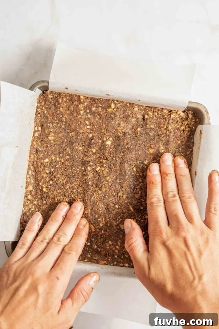 Two hands flattening energy bar dough evenly into a parchment paper-lined metal baking pan, illustrating the pressing technique.