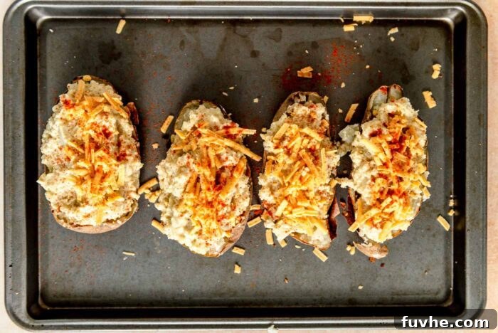 Four perfectly prepared vegan twice-baked potatoes, topped with a generous sprinkle of vegan cheese, arranged on a baking sheet, ready for their second bake.