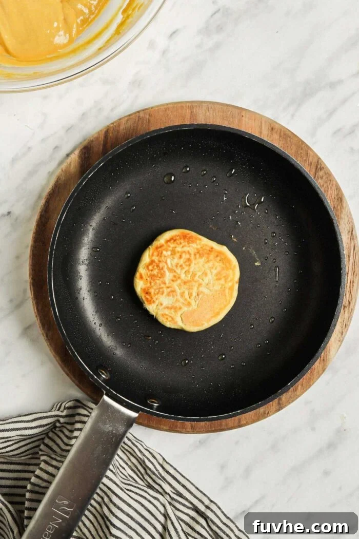 Savory Chickpea Griddle Cakes 6 A single, round garbanzo bean flour pancake sizzling and cooking in a hot, non-stick skillet. Its surface shows developing bubbles, indicating it's nearly ready to be flipped, while the edges appear slightly set.