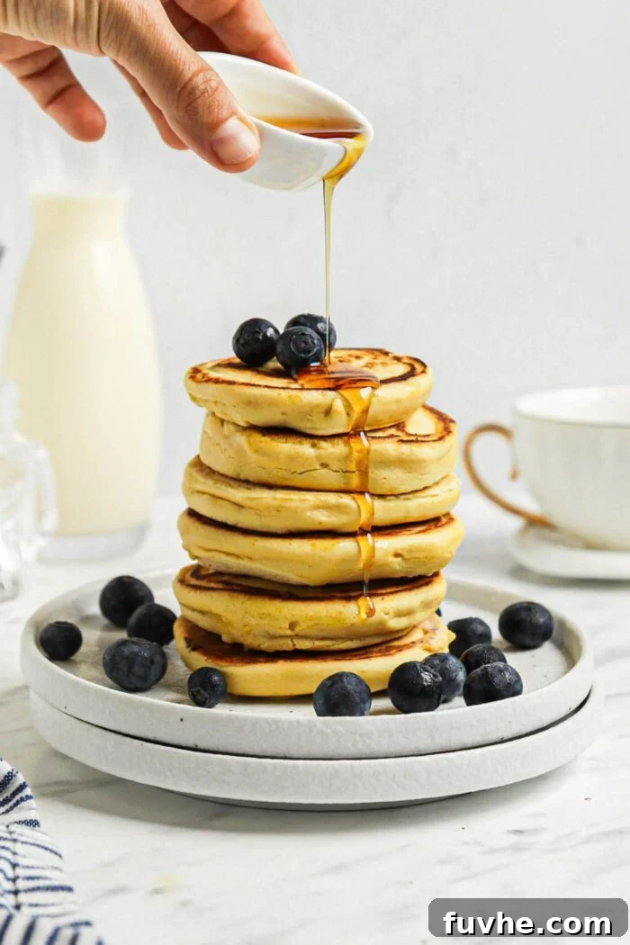 Savory Chickpea Griddle Cakes 7 A hand gently drizzles rich maple syrup from a small ceramic pitcher over a towering stack of freshly made chickpea pancakes, which are also adorned with vibrant blueberries, all resting on a white plate, ready to be enjoyed.