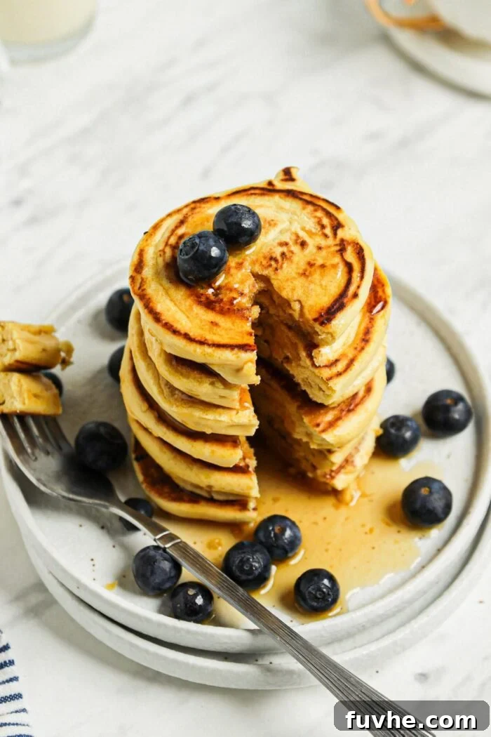 Savory Chickpea Griddle Cakes 8 A large, inviting stack of golden chickpea blueberry pancakes rests on a white plate, with a single slice neatly removed by a fork, showcasing the fluffy interior and embedded berries. The setting is clean and appetizing.