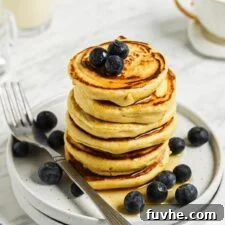 A perfectly arranged stack of 6 thick chickpea flour pancakes, beautifully adorned with fresh blueberries and a generous drizzle of maple syrup, presented on a small plate. The visual highlights the inviting texture and delicious toppings.