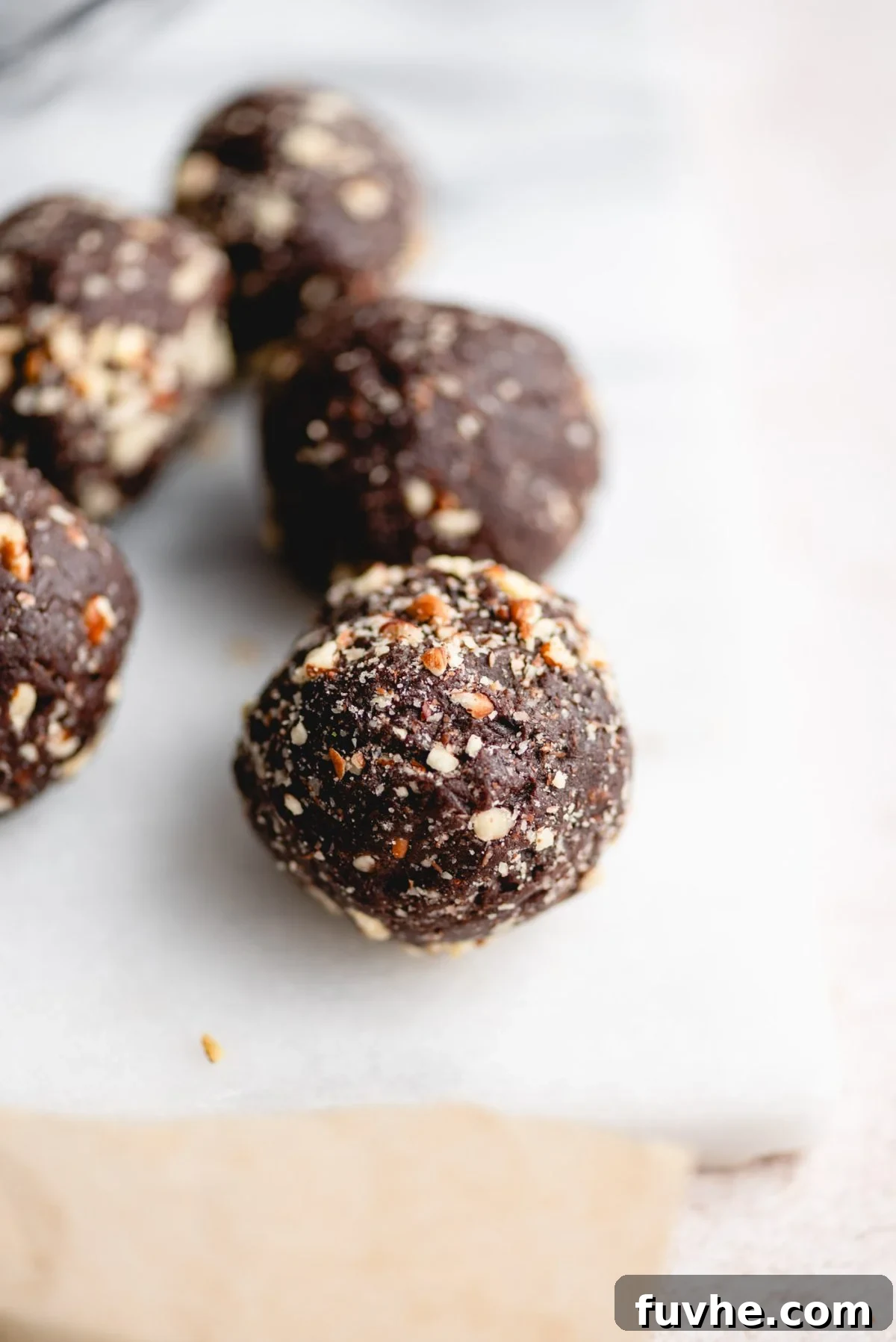 Chocolate Date Truffles 6 Several rich chocolate date balls, each beautifully coated in crushed pecans, are artfully arranged on a sleek marble cutting board, highlighting their enticing texture and gourmet appearance.