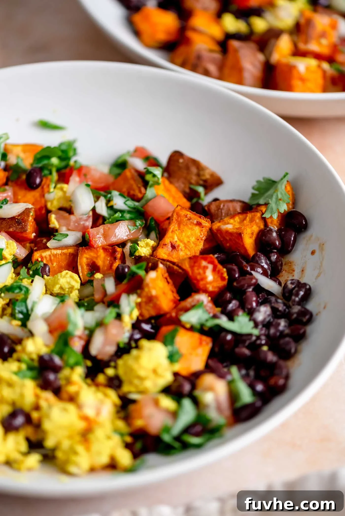 A beautifully assembled vegan breakfast burrito bowl featuring a golden tofu scramble, black beans, vibrant salsa, and roasted sweet potato cubes, garnished with fresh cilantro and a drizzle of sauce.