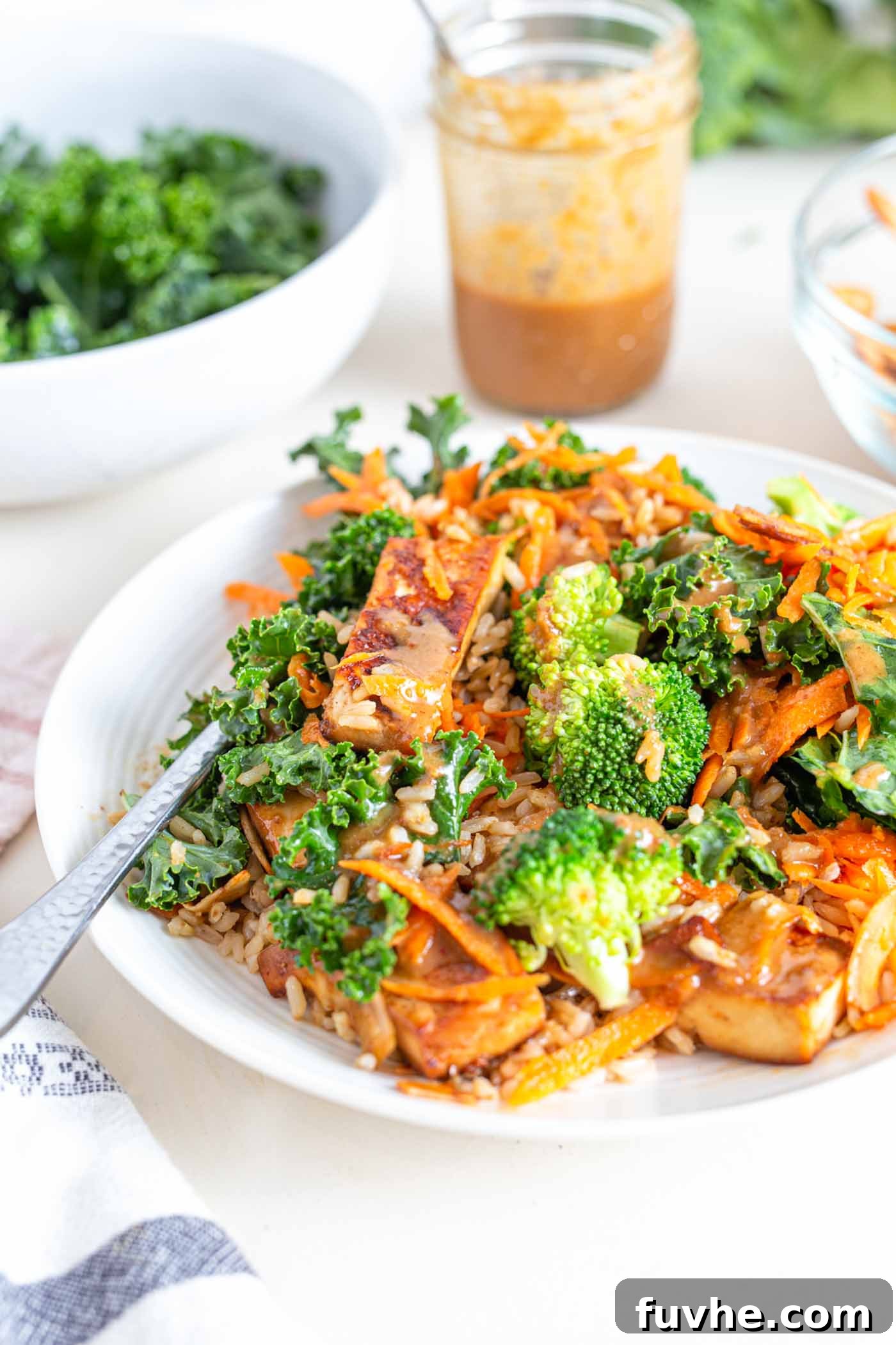 Green Harvest Bowl 2 A beautifully arranged bowl featuring golden baked tofu, bright green broccoli florets, shredded carrots, kale, and a drizzle of creamy almond butter sauce, garnished with crispy coconut bacon.