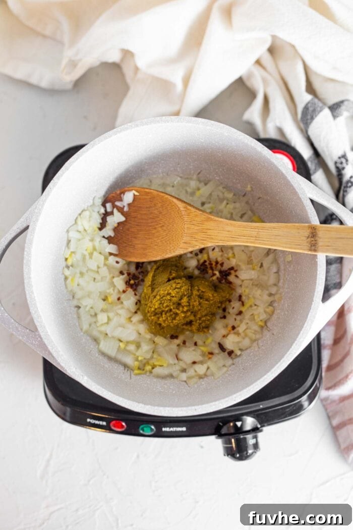 Fragrant Vegan Thai Green Curry 5 A wooden spoon actively stirring vibrant green curry paste into a pot with sautéed onions and garlic, preparing the flavor base.