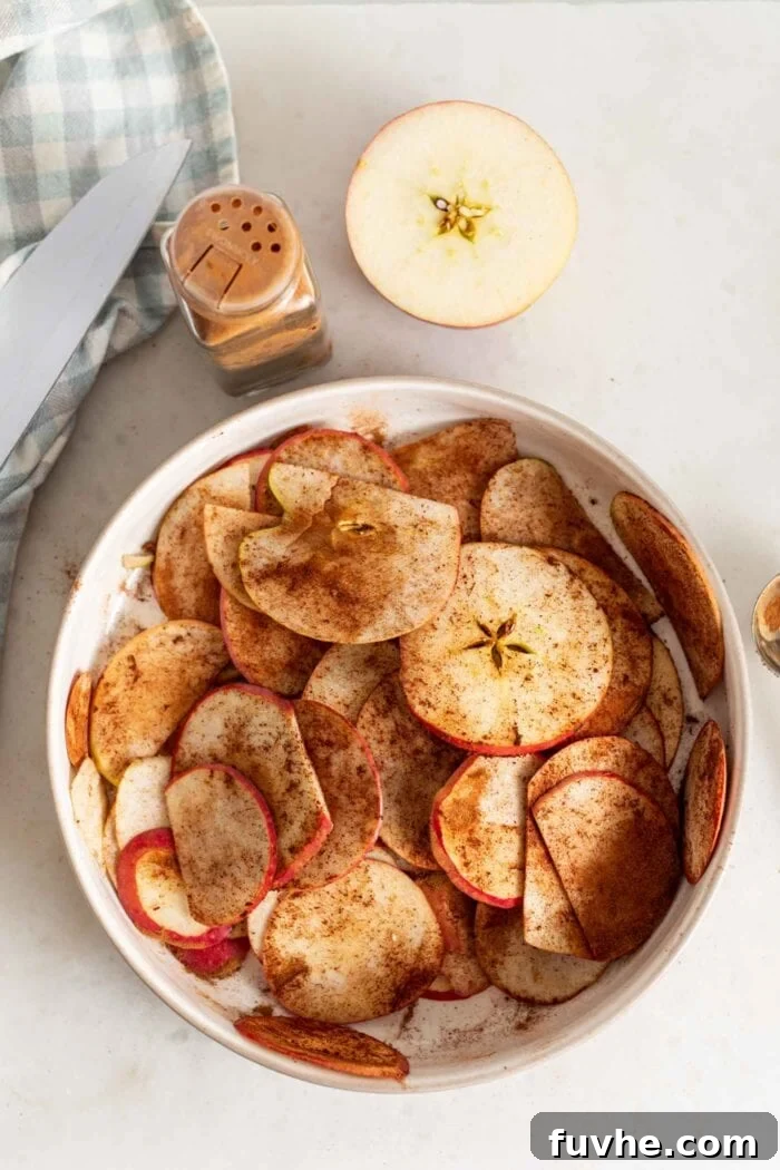 A vibrant close-up of thinly sliced apples, beautifully coated in a generous, even layer of cinnamon, resting in a glass bowl and ready for baking. The cinnamon adheres perfectly to the moist apple surfaces.