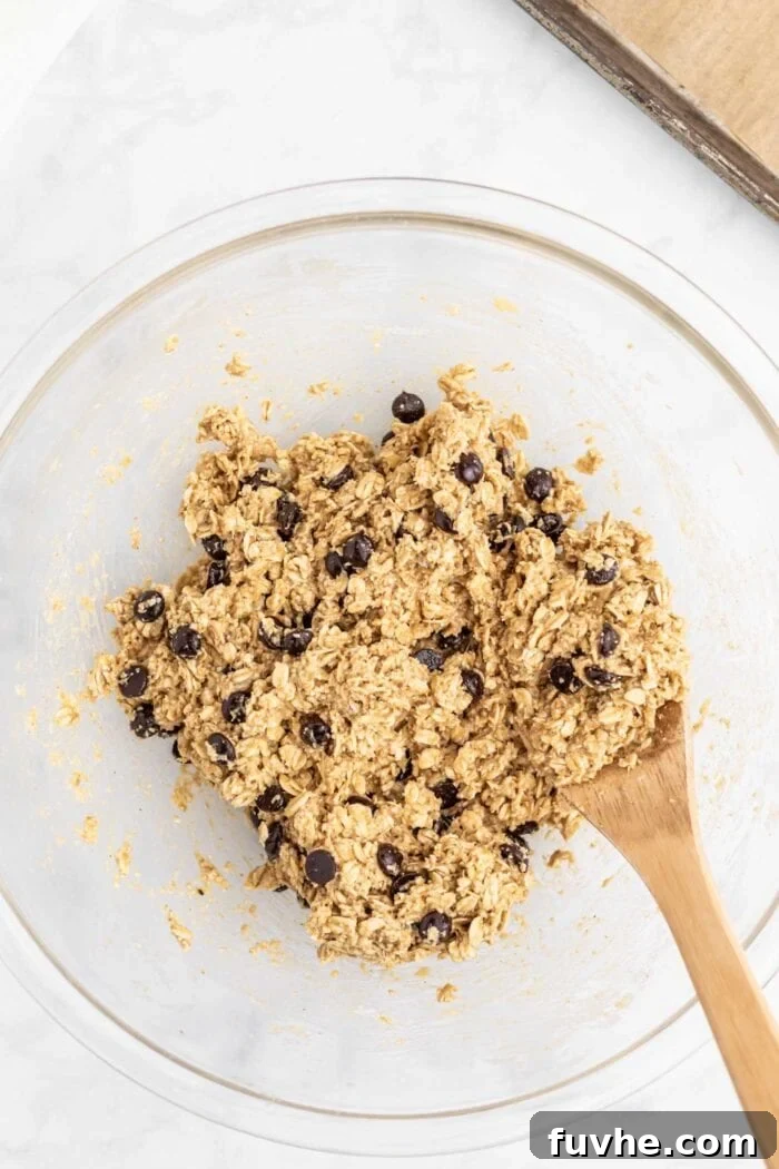Irresistible Vegan Peanut Butter Oatmeal Cookies 7 A rich, sticky oatmeal chocolate chip cookie dough in a glass mixing bowl, featuring visible oats and chocolate chips, mixed with a wooden spoon.