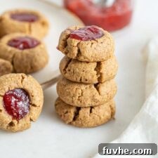 A stack of four golden-brown thumbprint cookies, each with a bright jam center, next to a larger plate of cookies, symbolizing a finished batch.