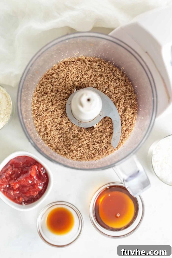 Finely blended pecans in a food processor, flanked by small bowls of vibrant jam, golden maple syrup, and clear vanilla extract, illustrating the initial steps of cookie preparation.