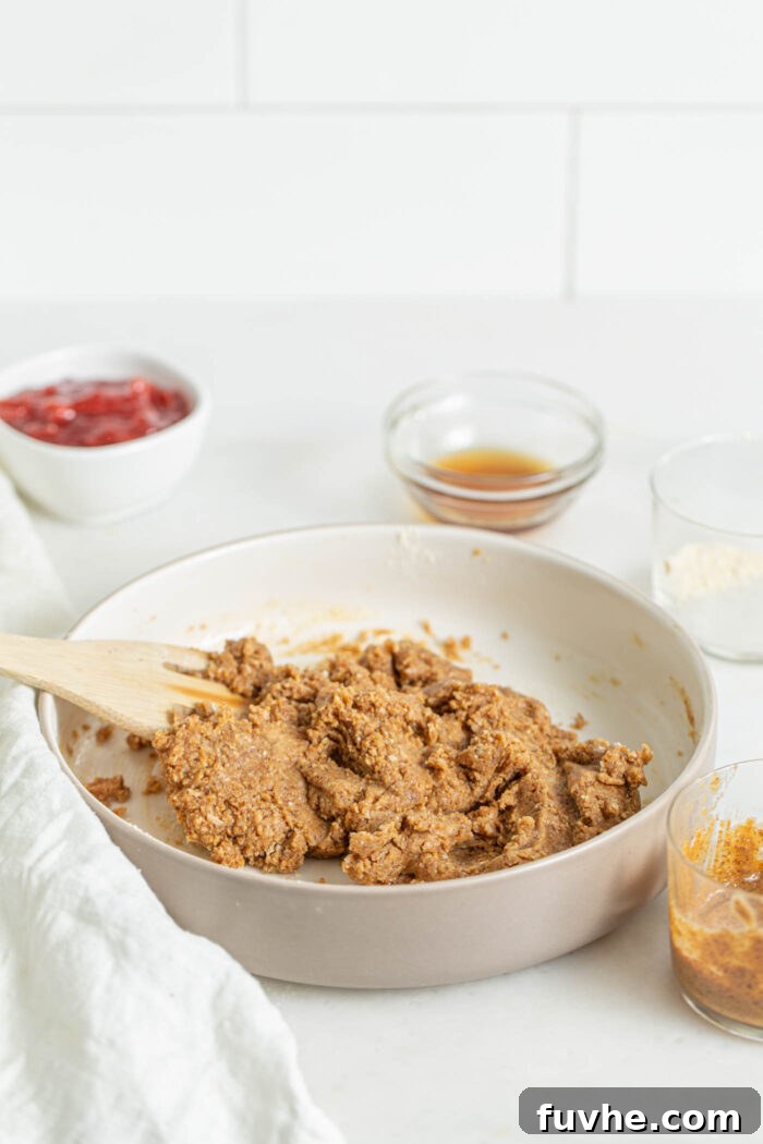 Raw cookie dough resting in a mixing bowl, accompanied by small dishes containing jam, vanilla extract, and almond butter, ready for shaping.