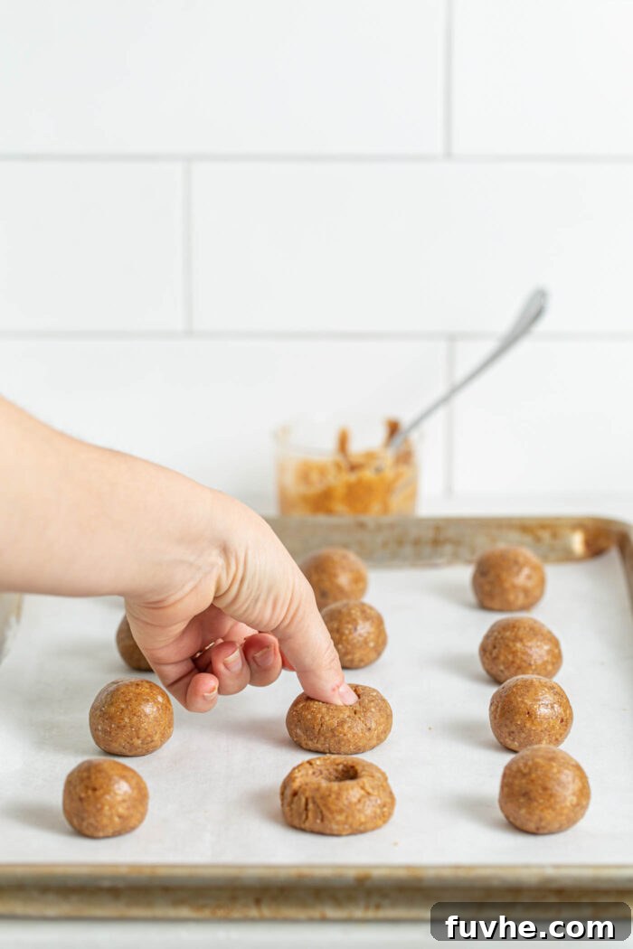 A hand gently presses a thumb into a raw cookie dough ball on a parchment paper-lined baking sheet, creating the signature thumbprint well.