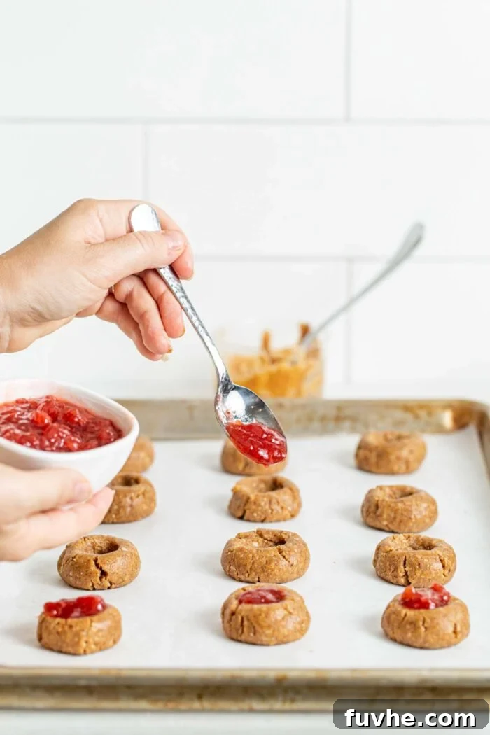 Jam being carefully spooned into the thumbprint indentations of raw cookie dough balls on a baking tray, showcasing the vibrant filling process.