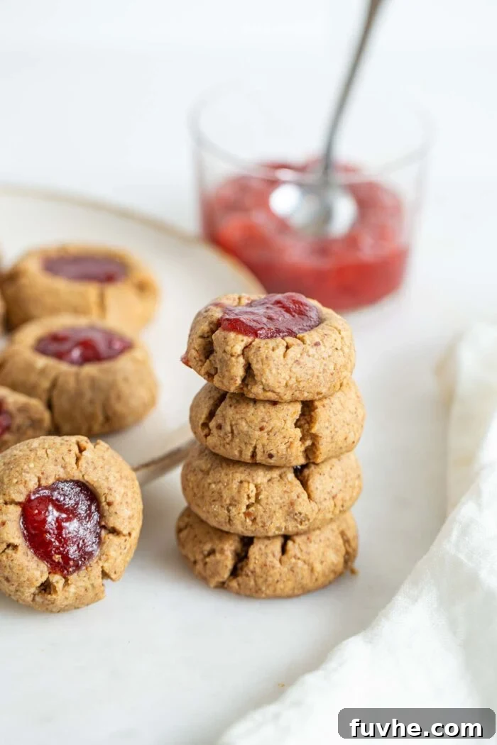 A beautifully arranged stack of four baked thumbprint cookies, sitting beside a plate full of more cookies, highlighting their golden edges and vibrant jam centers.