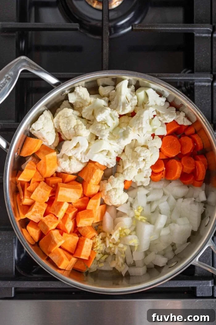 Sweet potato, cauliflower, carrot, onion, garlic, and ginger cooking in a stock pot over medium heat on a stovetop, building the aromatic base for the soup.