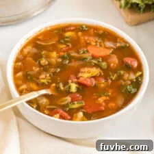 A close-up of a rustic bowl of tomato vegetable soup, featuring hearty lentils and vibrant kale, with a spoon resting in it.