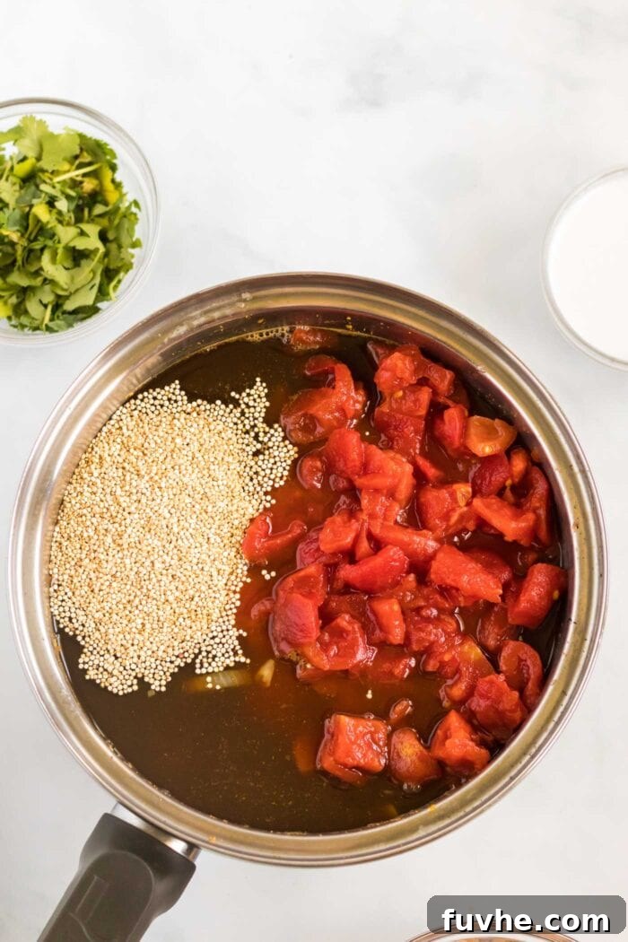A close-up view of diced canned tomatoes and uncooked quinoa added to the gently simmering vegetable broth in a large soup pot, ready to begin cooking.