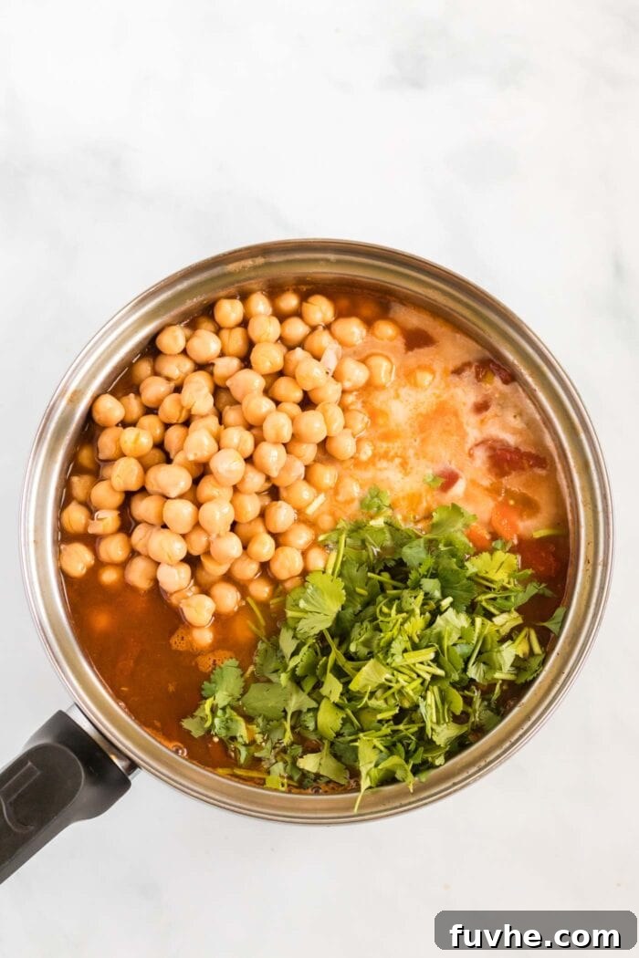 Plump chickpeas, vibrant fresh cilantro, and creamy coconut milk being gently stirred into the fragrant tomato broth in a soup pot, completing the rich and aromatic stew.