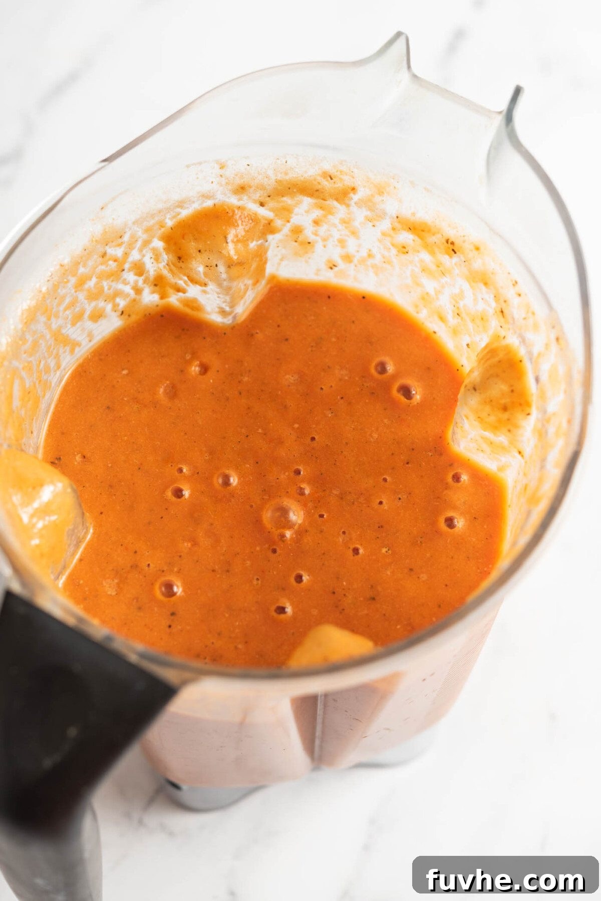 A high-speed blender swirling a vibrant, creamy mixture of roasted red pepper and tomato soup, ready for heating and serving.