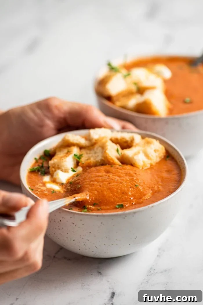 A hand gently dips a spoon into a comforting bowl of roasted red pepper tomato soup, with a second bowl softly blurred in the background, inviting a warm embrace.
