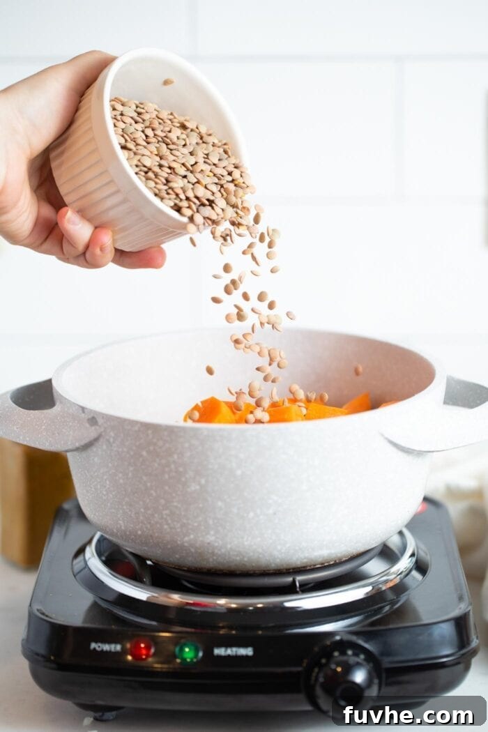 Golden Squash Lentil Pot 5 A hand carefully dumping a generous amount of lentils into a large pot filled with an assortment of chopped vegetables and broth.
