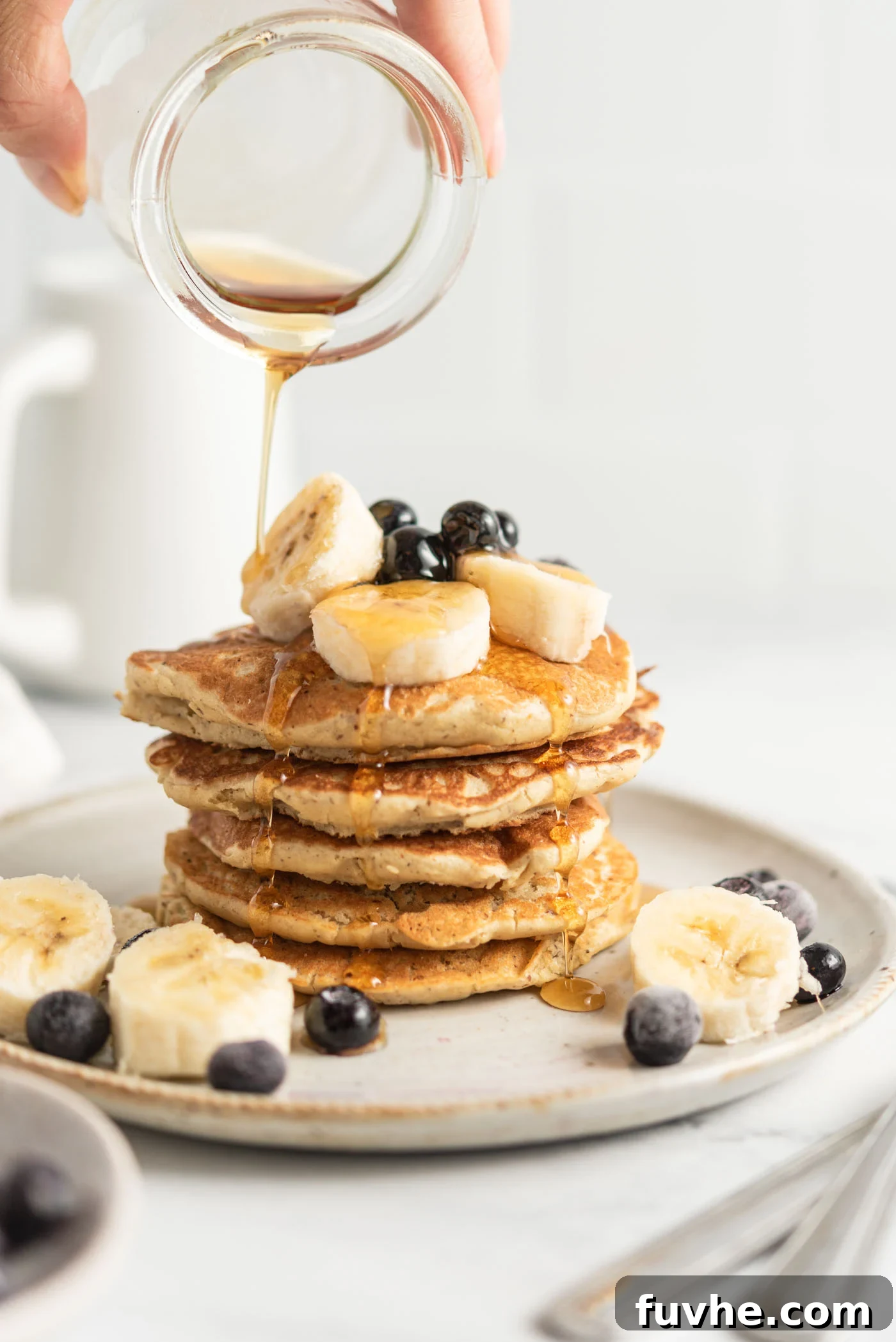 Protein-Packed Vegan Quinoa Pancakes 2 Hand pouring maple syrup from a small jar onto a stack of quinoa flour pancakes topped with banana and blueberry.