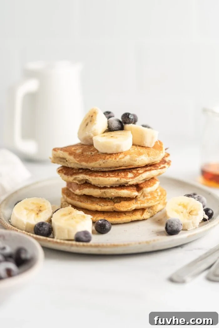 Protein-Packed Vegan Quinoa Pancakes 9 Stack of quinoa flour pancakes topped with blueberries and banana on a small plate. There's a small jug in the background and a small jar of maple syrup off to the side.