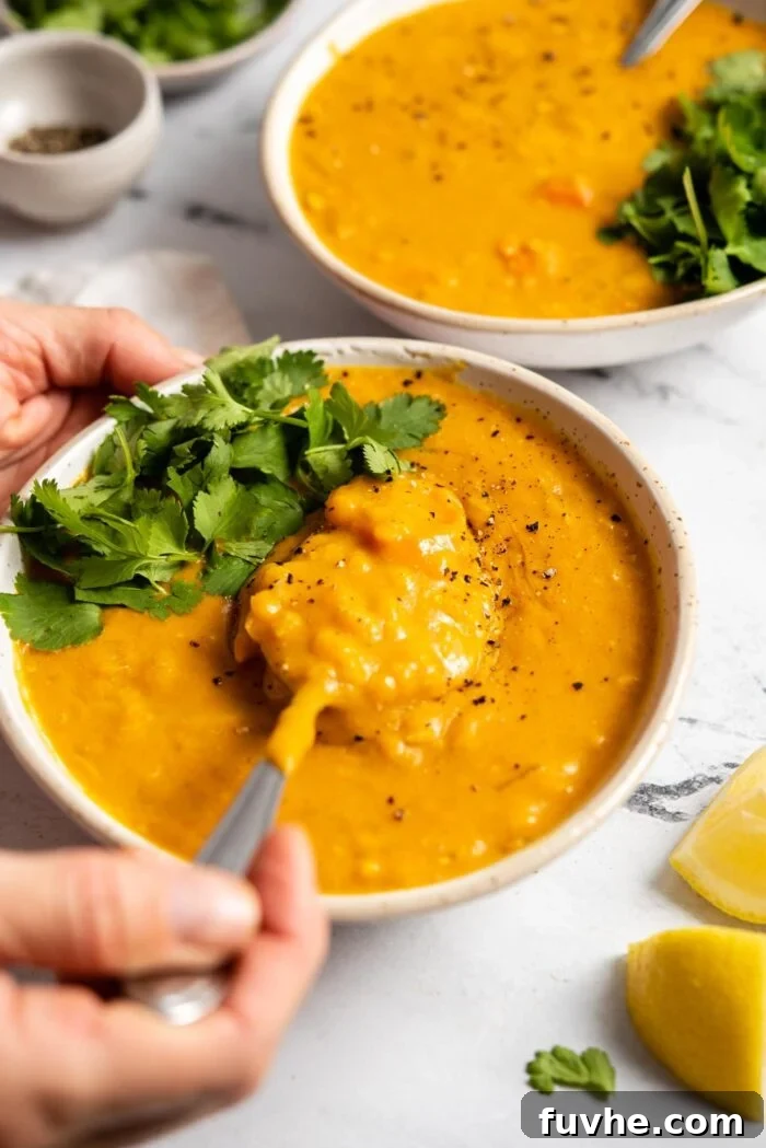A hand skillfully uses a spoon to lift a spoonful of vibrant lemon lentil soup, beautifully topped with fresh cilantro, from a cozy bowl. Another bowl of the delicious soup is subtly blurred in the background, completing the inviting scene.