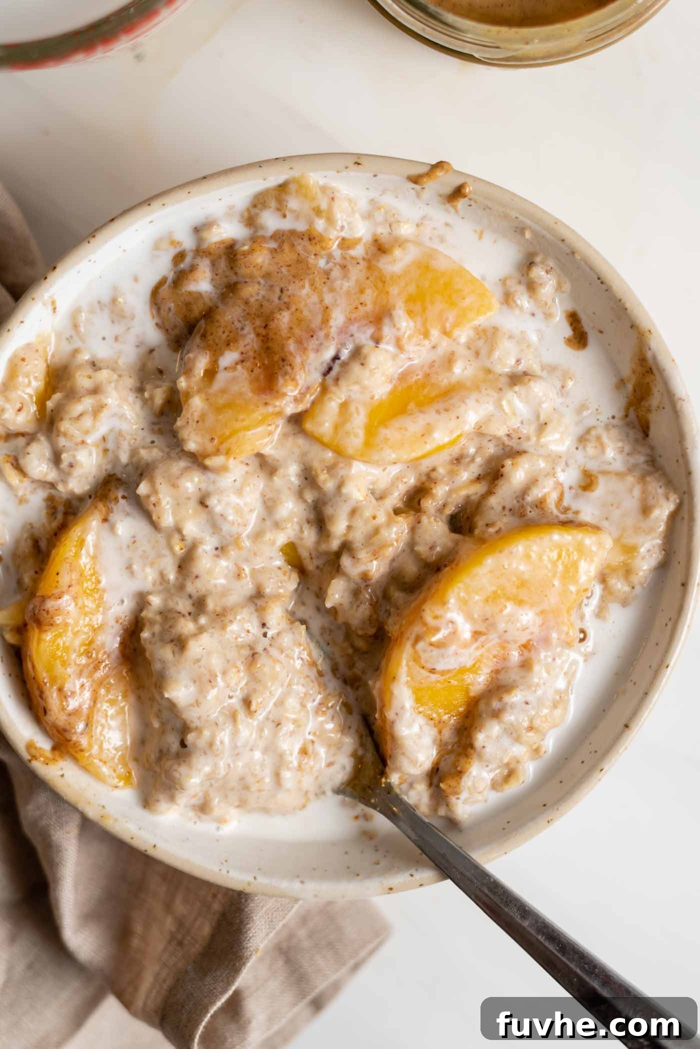 Velvety Vegan Peach Dream Bowl 2 A bowl of peaches and cream oatmeal topped with coconut milk and almond butter. The bowl sits on a dish cloth and there's a spoon in the bowl.