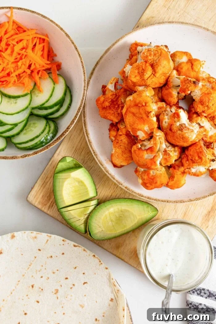 Tangy Buffalo Cauliflower Wraps 8 A wooden cutting board with a bowl of buffalo cauliflower, alongside neatly sliced avocado, cucumber, and grated carrot, ready for wrap assembly.
