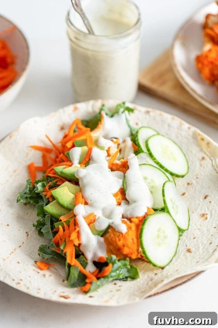 Tangy Buffalo Cauliflower Wraps 9 An open tortilla wrap filled with cucumber, avocado, buffalo cauliflower, carrot, and a generous drizzle of creamy sauce, ready to be rolled.