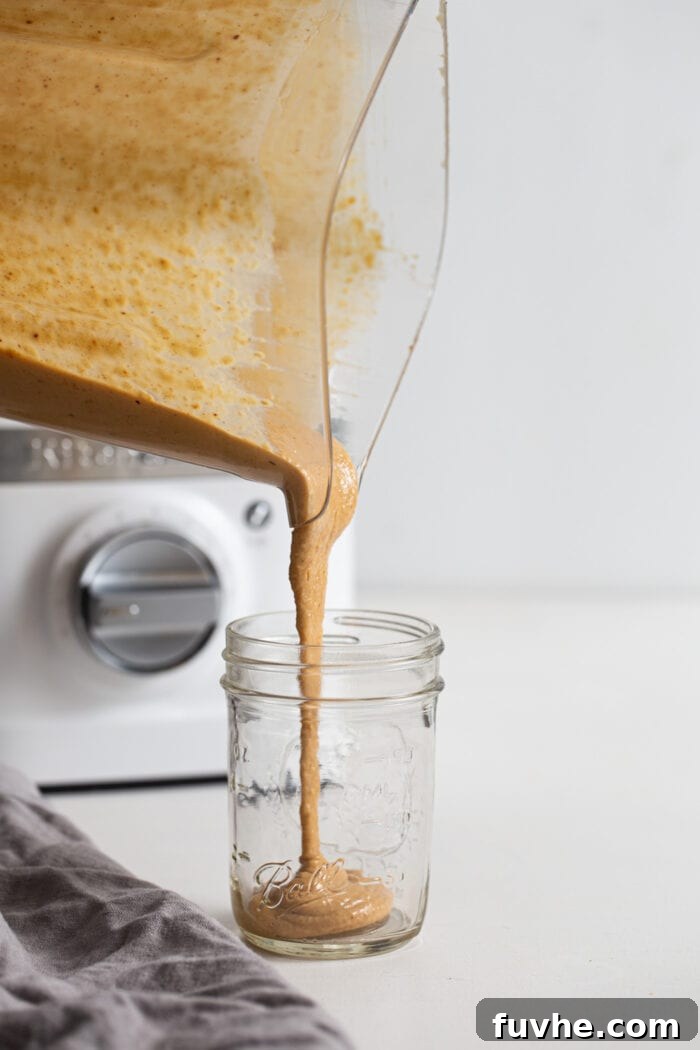 A blender pouring thick, creamy peanut sauce into a glass jar.