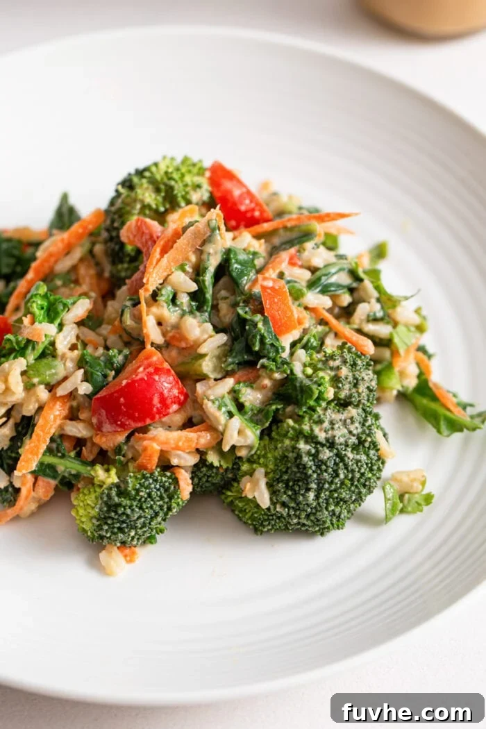 A close-up shot of a vibrant broccoli, carrot, and bell pepper salad generously coated with creamy peanut sauce on a white plate.