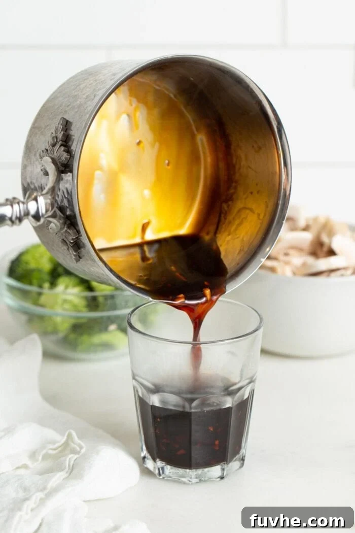 Rich, homemade teriyaki sauce being poured from a small saucepan into a clear glass measuring cup, showcasing its thick and glossy texture.