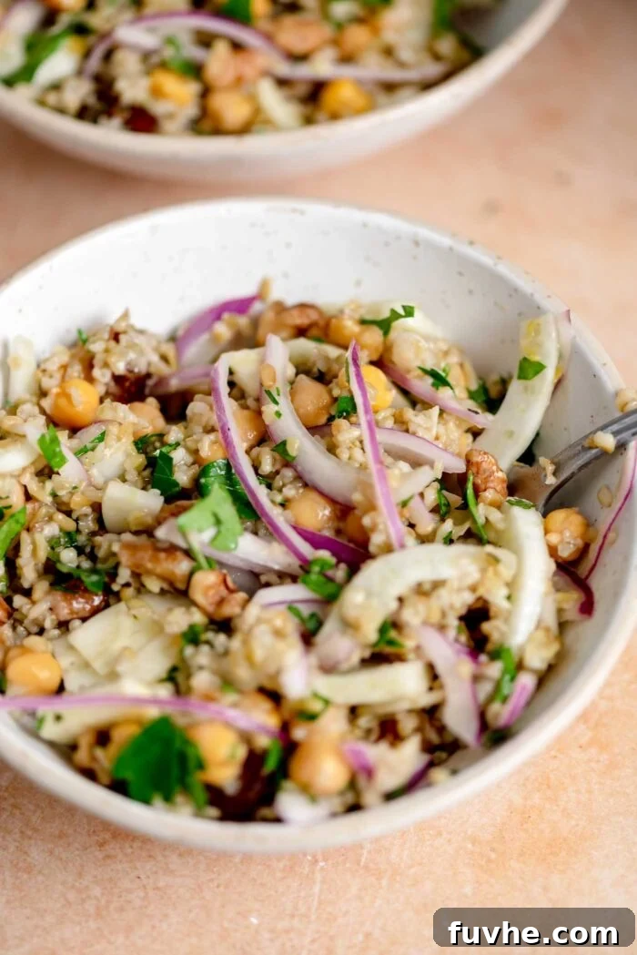 Close-up of a serving bowl filled with Freekeh Salad, featuring visible red onion, sliced fennel, chickpeas, sweet dates, walnuts, and fresh herbs, ready to be enjoyed.